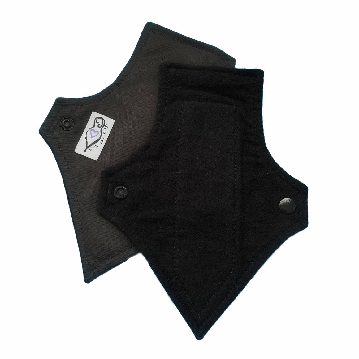 Two reusable menstrual pads with a squared top and tapered bottom shape, against a white background. The Pad on top is made from black flannel fabric. The Pad underneath is made from black PUL fabric and has a white 'Scarlet Eve' label.