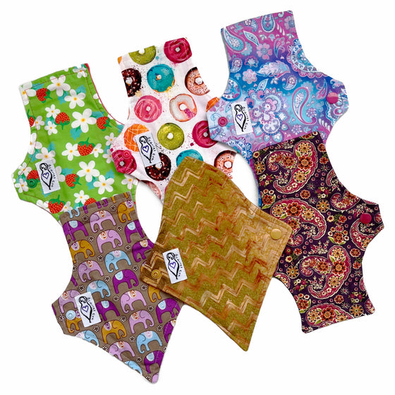 A set of six assorted panty liners with various colorful patterns, against a white background. Some Pads are squared at the top and one is curved. Some Pads are flared at the bottom and some are tapered.