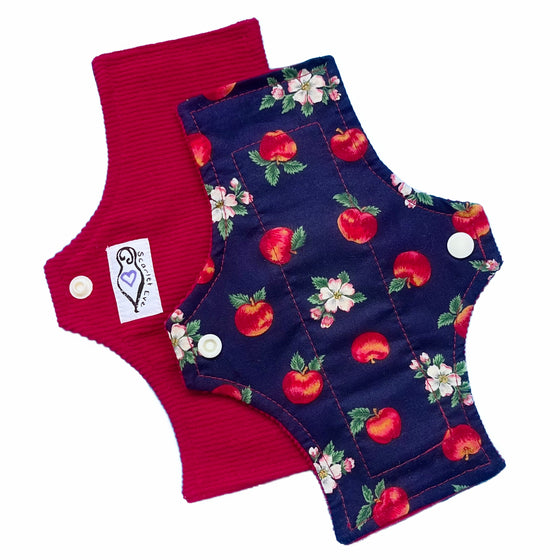 Two resusable panty liners against a white background. The pad on top is made from navy blue cotton fabric with a red apples and white flowers pattern. The Pad underneath is made from dark red corduroy and has a white 'Scarlet Eve' label.