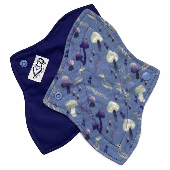 Two reusable menstrual pads with a contoured shape against a white background. The pad on top is made from blue cotton flannel with purple and grey mushrooms. The pad on the bottom is made from purple PUL fabric and has a white "Scarlet Eve" label.