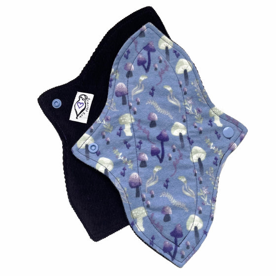 Two reusable menstrual pads with a contoured shape against a white background. The pad on top is made from blue cotton flannel with purple and grey mushrooms. The pad on the bottom is made from dark purple cotton corduroy fabric and has a white "Scarlet Eve" label.