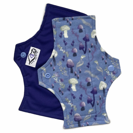 Two reusable menstrual pads with squared top and flared bottom shape against a white background. The pad on top is made from blue cotton flannel with purple and grey mushrooms. The pad on the bottom is made from purple PUL fabric and has a white "Scarlet Eve" label.