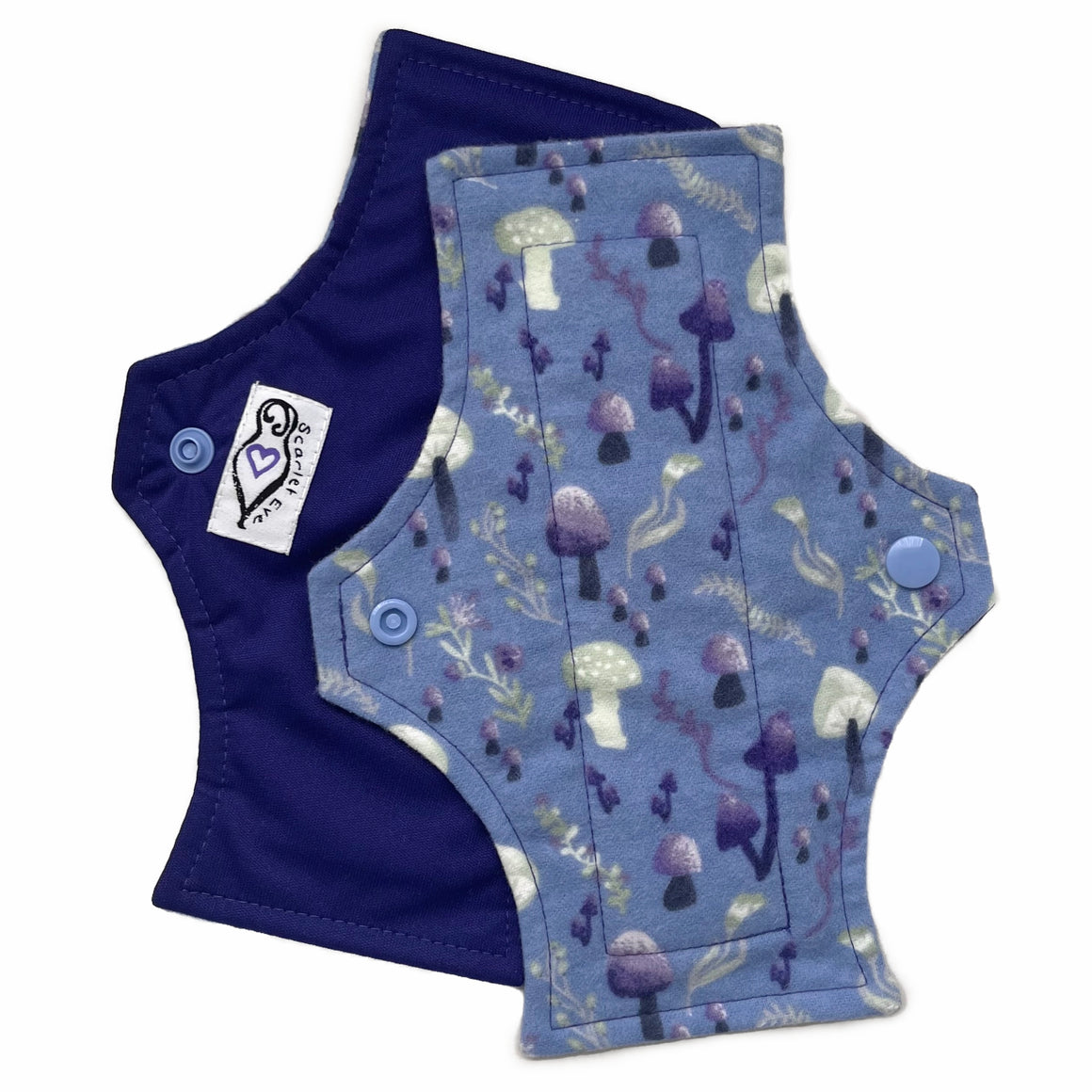 Two reusable menstrual pads with squared top and flared bottom shape against a white background. The pad on top is made from blue cotton flannel with purple and grey mushrooms. The pad on the bottom is made from purple PUL fabric and has a white "Scarlet Eve" label.