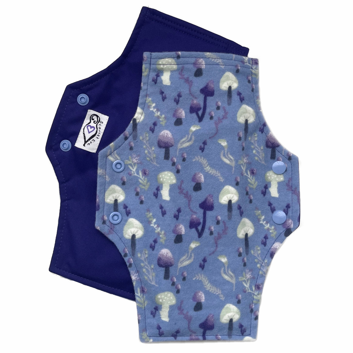 Two reusable menstrual pads with squared top and flared bottom shape against a white background. The pad on top is made from blue cotton flannel with purple and grey mushrooms. The pad on the bottom is made from purple PUL fabric and has a white "Scarlet Eve" label.