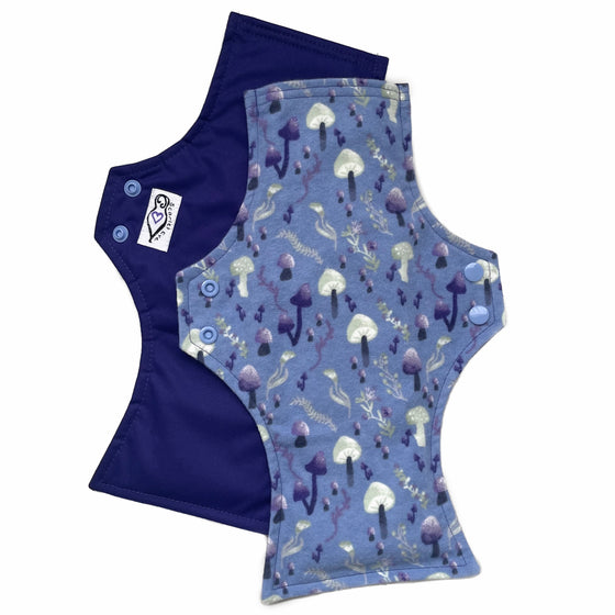 Two reusable menstrual pads with squared top and flared bottom shape against a white background. The pad on top is made from blue cotton flannel with purple and grey mushrooms. The pad on the bottom is made from purple PUL fabric and has a white "Scarlet Eve" label.