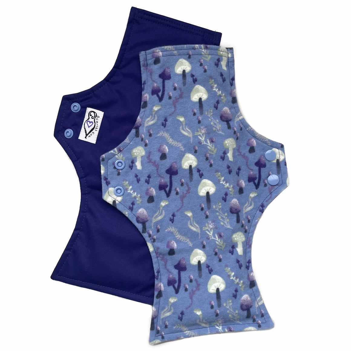 Two reusable menstrual pads with squared top and flared bottom shape against a white background. The pad on top is made from blue cotton flannel with purple and grey mushrooms. The pad on the bottom is made from purple PUL fabric and has a white "Scarlet Eve" label.