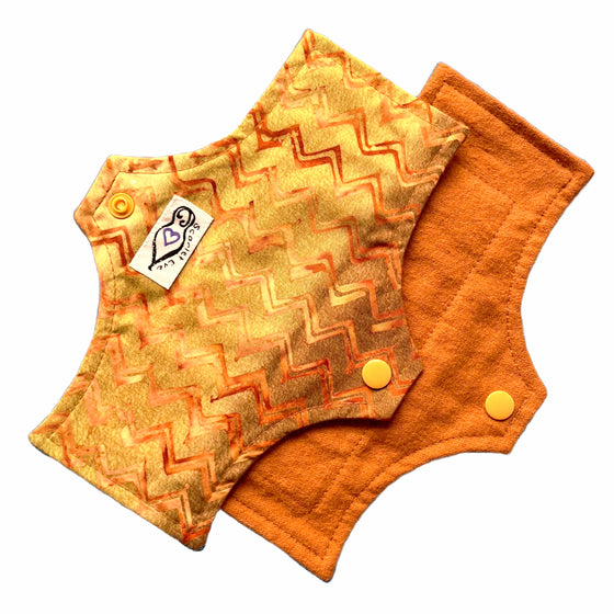 Two reusable panty liners against a white background. The Pad on top is made from golden brown chevron cotton fabric with a white 'Scarlet Eve' label. The Pad underneath is made from caramel brown flannel fabric.