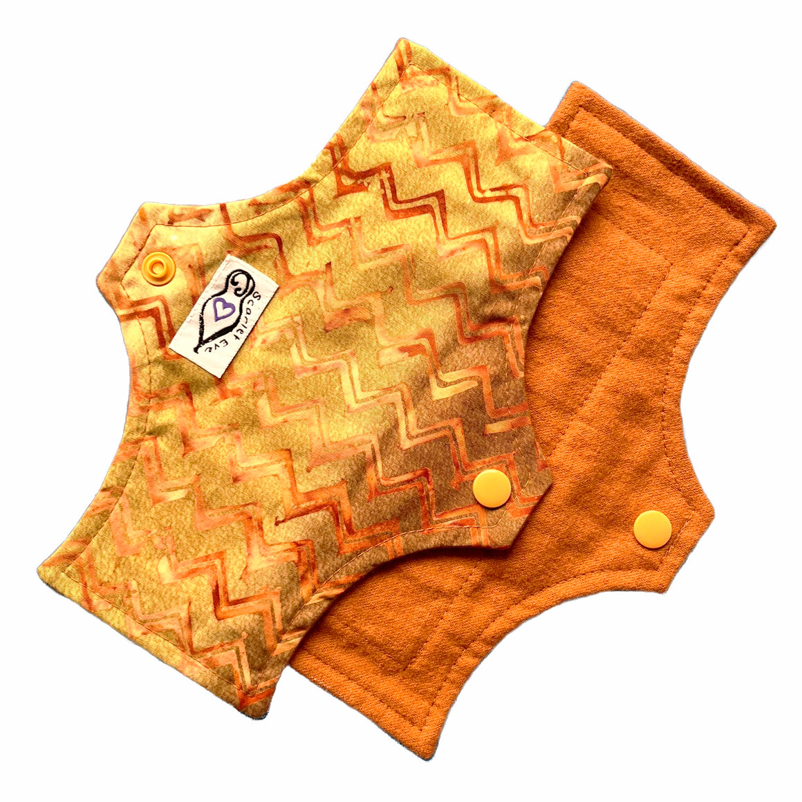 Two reusable panty liners against a white background. The Pad on top is made from golden brown chevron cotton fabric with a white 'Scarlet Eve' label. The Pad underneath is made from caramel brown flannel fabric.