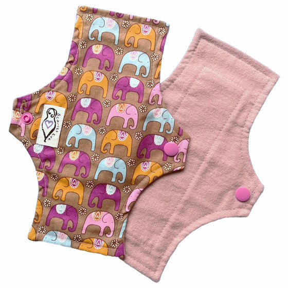 Two reusable panty liners against a white background. The Pad on top is made from brown cotton with orange, purple and blue elephants, and a white 'Scarlet Eve' label. The Pad underneath is made from lilac purple flannel fabric.