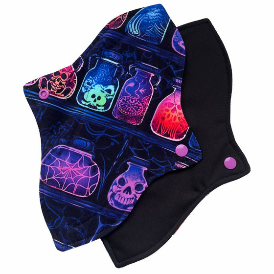 Two reusable menstrual pads with a contoured shape against a white background. The pad on top is made from black cotton Lycra with potion bottles in neon colours. The pad on the bottom is made from black PUL fabric and has a white "Scarlet Eve" label.