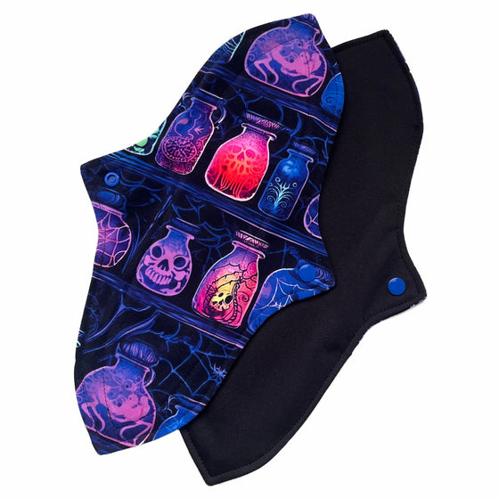 Two reusable menstrual pads with a contoured shape against a white background. The pad on top is made from black cotton Lycra with potion bottles in neon colours. The pad on the bottom is made from black PUL fabric and has a white "Scarlet Eve" label.