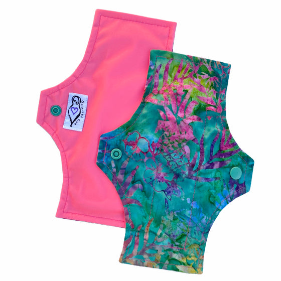 Two reusable menstrual pads with a squared top and flared bottom shape, against a white background. The pad on top is made from jade green batik cotton fabric with bright yellow, pink, blue and purple pineapple print. The pad on the bottom is made from coral pink PUL fabric, and has a white "Scarlet Eve" label.