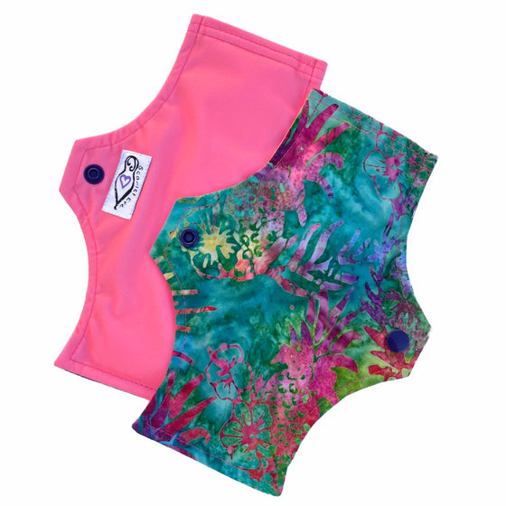 Two reusable menstrual pads with a squared top and flared bottom shape, against a white background. The pad on top is made from jade green batik cotton fabric with bright yellow, pink, blue and purple pineapple print. The pad on the bottom is made from coral pink PUL fabric, and has a white "Scarlet Eve" label.