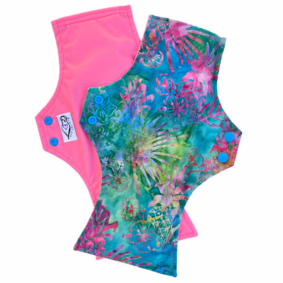 Two reusable menstrual pads with a squared top and flared bottom shape, against a white background. The pad on top is made from jade green batik cotton fabric with bright yellow, pink, blue and purple pineapple print. The pad on the bottom is made from coral pink PUL fabric, and has a white "Scarlet Eve" label.