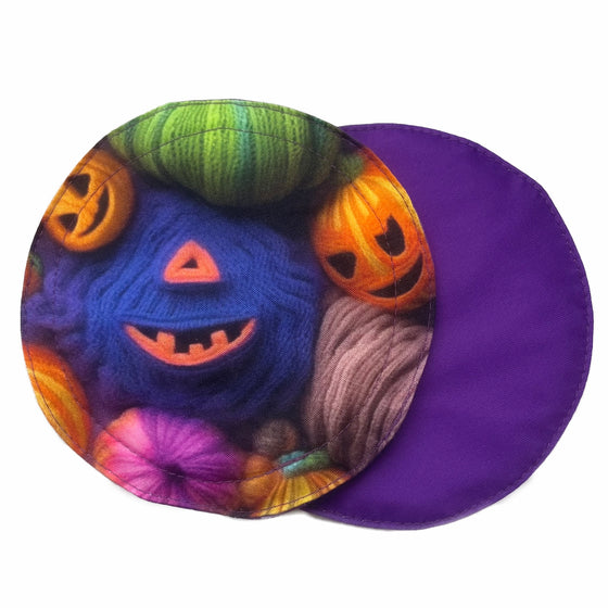 Two reusable breast pads against a white background. The breast pad on top is made from woven cotton with orange and purple pumpkins made of yarn. The breast pad on the bottom is made from purple PUL fabric.