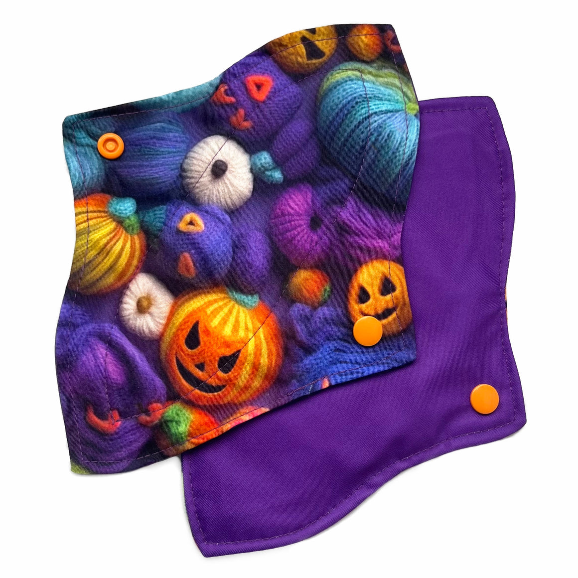 Two reusable menstrual pads with a contoured shape against a white background. The pad on top is made from purple woven cotton with orange, purple, green and blue pumpkins made from yarn. The pad on the bottom is made from purple PUL fabric and has a white "Scarlet Eve" label.