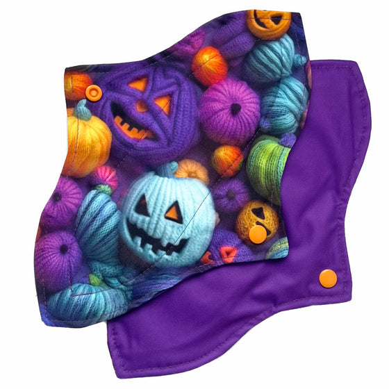 Two reusable menstrual pads with a contoured shape against a white background. The pad on top is made from purple woven cotton with orange, purple, green and blue pumpkins made from yarn. The pad on the bottom is made from purple PUL fabric and has a white "Scarlet Eve" label.