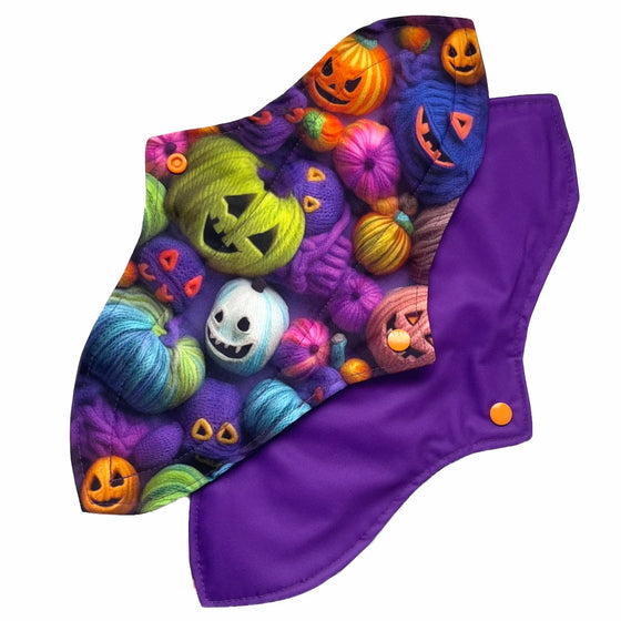 Two reusable menstrual pads with a contoured shape against a white background. The pad on top is made from purple woven cotton with orange, purple, green and blue pumpkins made from yarn. The pad on the bottom is made from purple PUL fabric and has a white "Scarlet Eve" label.