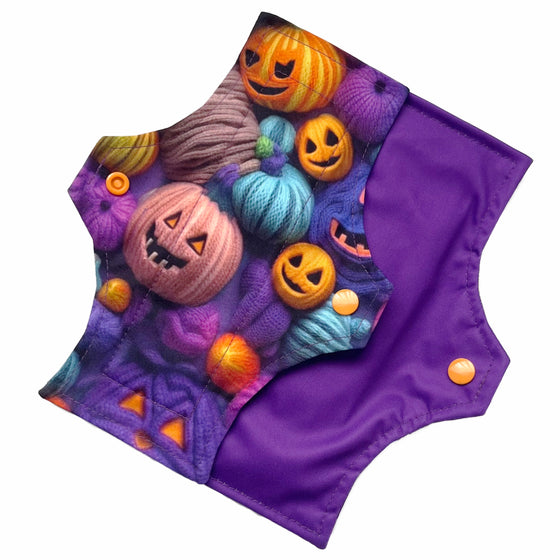 Two reusable menstrual pads with squared top and flared bottom shape against a white background. The pad on top is made from purple woven cotton with orange, purple, green and blue pumpkins made from yarn. The pad on the bottom is made from purple PUL fabric and has a white "Scarlet Eve" label.
