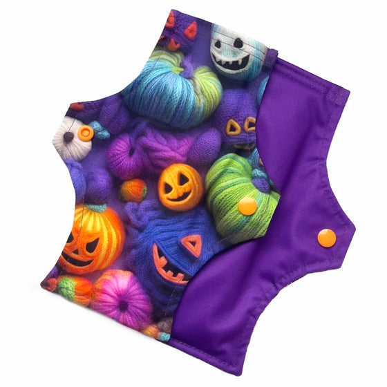 Two reusable menstrual pads with squared top and flared bottom shape against a white background. The pad on top is made from purple woven cotton with orange, purple, green and blue pumpkins made from yarn. The pad on the bottom is made from purple PUL fabric and has a white "Scarlet Eve" label.