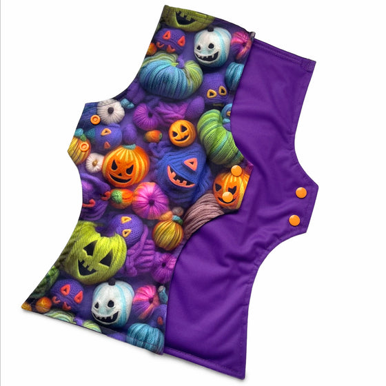 Two reusable menstrual pads with squared top and flared bottom shape against a white background. The pad on top is made from purple woven cotton with orange, purple, green and blue pumpkins made from yarn. The pad on the bottom is made from purple PUL fabric and has a white "Scarlet Eve" label.