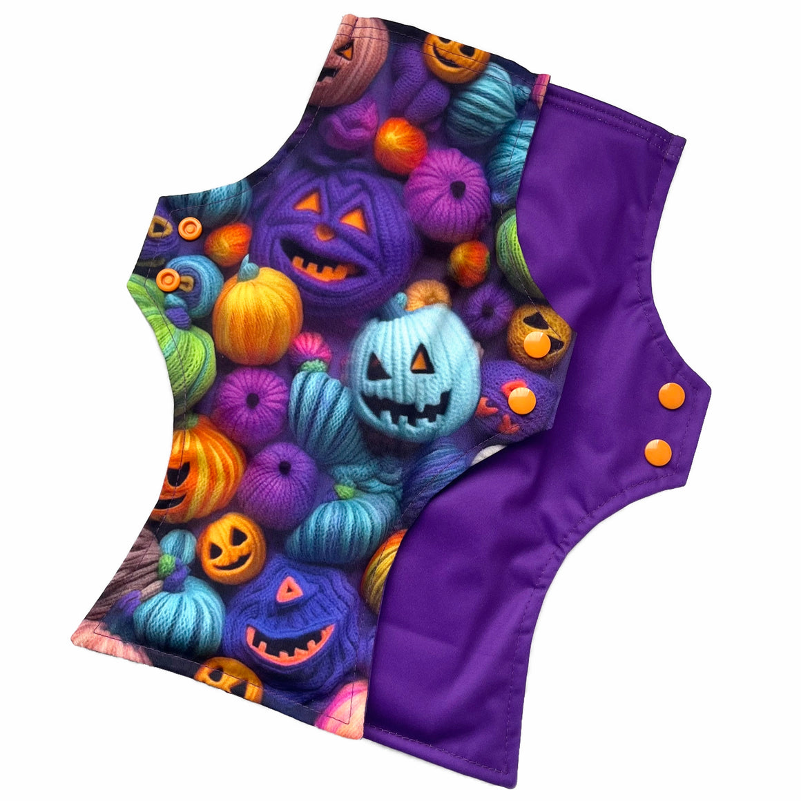 Two reusable menstrual pads with squared top and flared bottom shape against a white background. The pad on top is made from purple woven cotton with orange, purple, green and blue pumpkins made from yarn. The pad on the bottom is made from purple PUL fabric and has a white "Scarlet Eve" label.