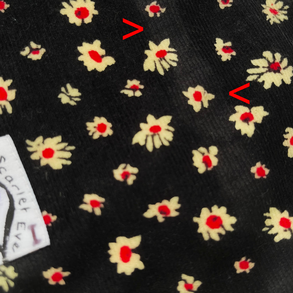 Close up of black corduroy fabric with white and red daisies. Red arrows point to a strip of lighter coloured corduroy running vertically along the fabric.