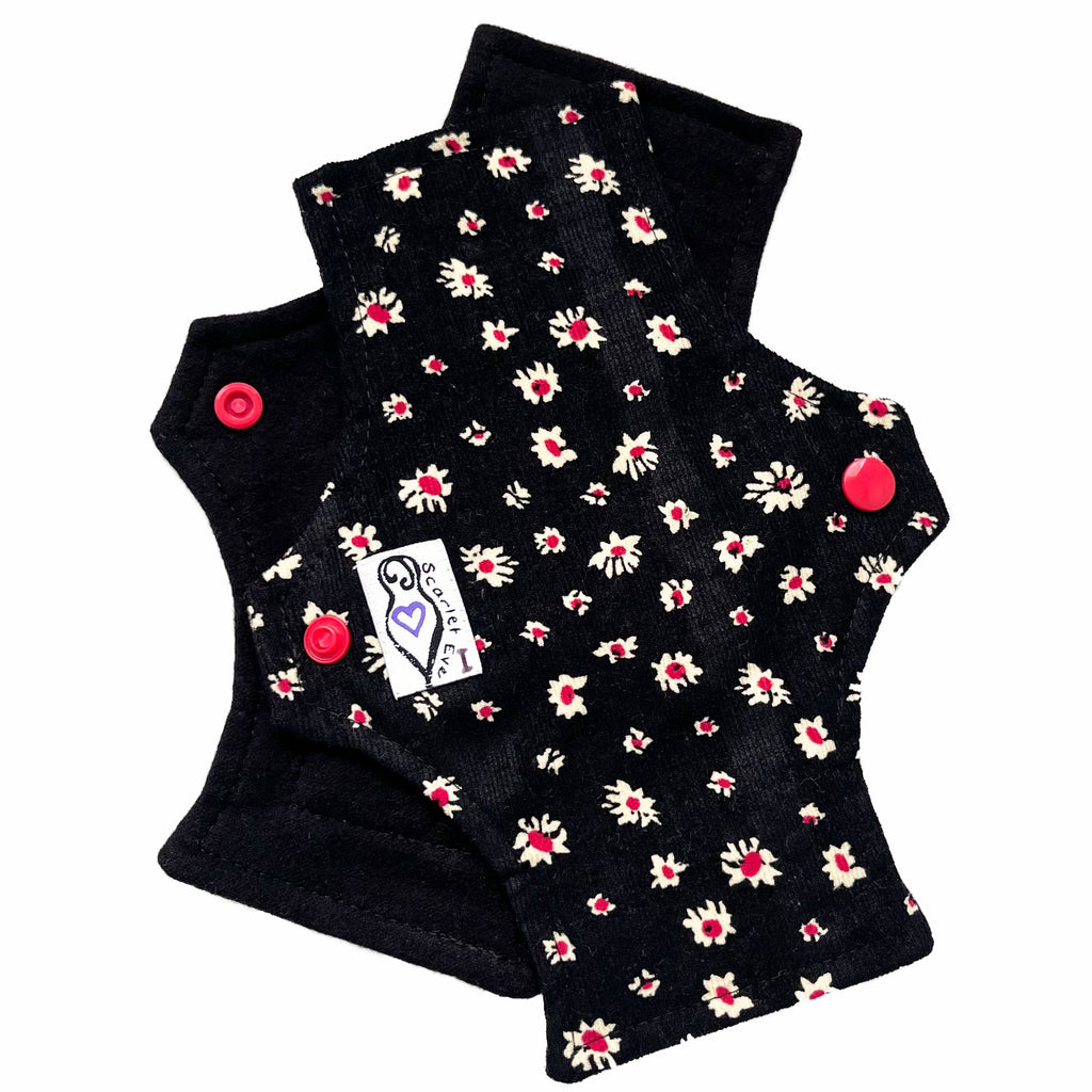 Two reusable panty liners against a white background. The pad on top is made from white and red daisies on black corduroy, and has a white 'Scarlet Eve' label with an I on it. The pad on the bottom is made from black flannel fabric.