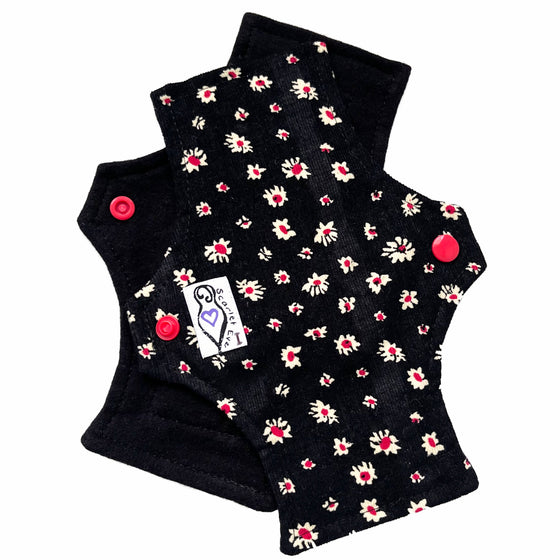 Two reusable panty liners against a white background. The pad on top is made from white and red daisies on black corduroy, and has a white 'Scarlet Eve' label with an I on it. The pad on the bottom is made from black flannel fabric.