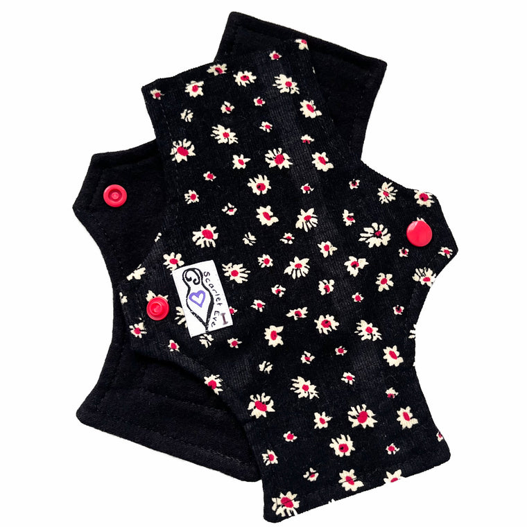 Two reusable panty liners against a white background. The pad on top is made from white and red daisies on black corduroy, and has a white 'Scarlet Eve' label with an I on it. The pad on the bottom is made from black flannel fabric.