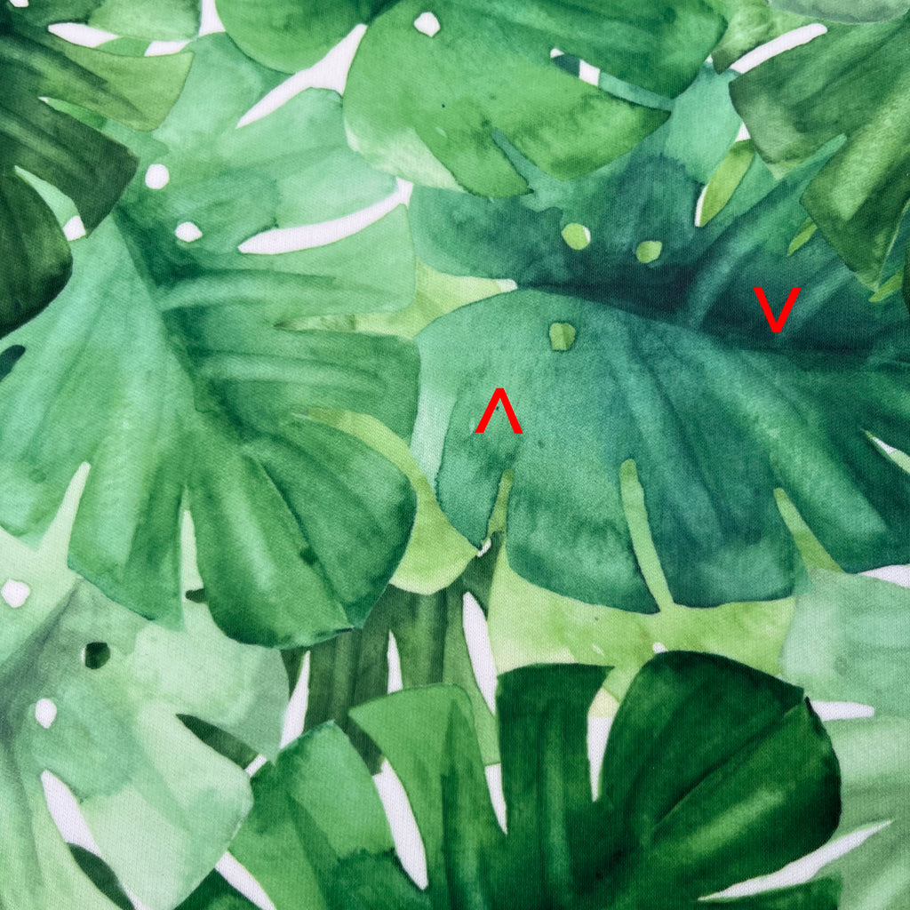 A close up of white PUL fabric with large monstera leaves in various shads of green. Red arrows point to indented fault lines running across the fabric.
