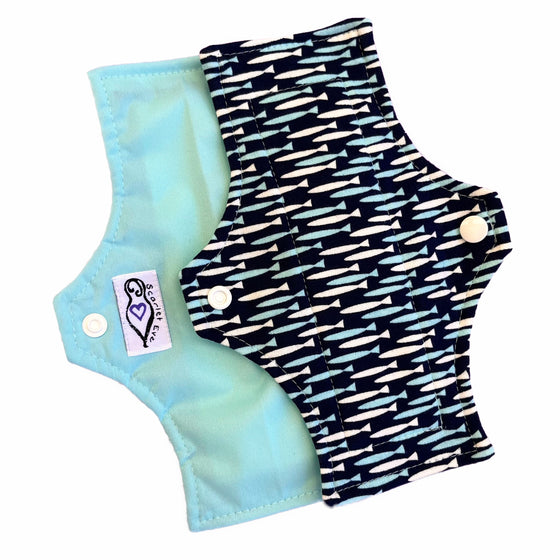 Two reusable menstrual pads with squared top and flared bottom shape, against a white background. The pad on top is made from navy jersey fabric with small turquoise and white fish print. The pad on the bottom is made from turquoise PUL fabric, and has a white "Scarlet Eve" label.