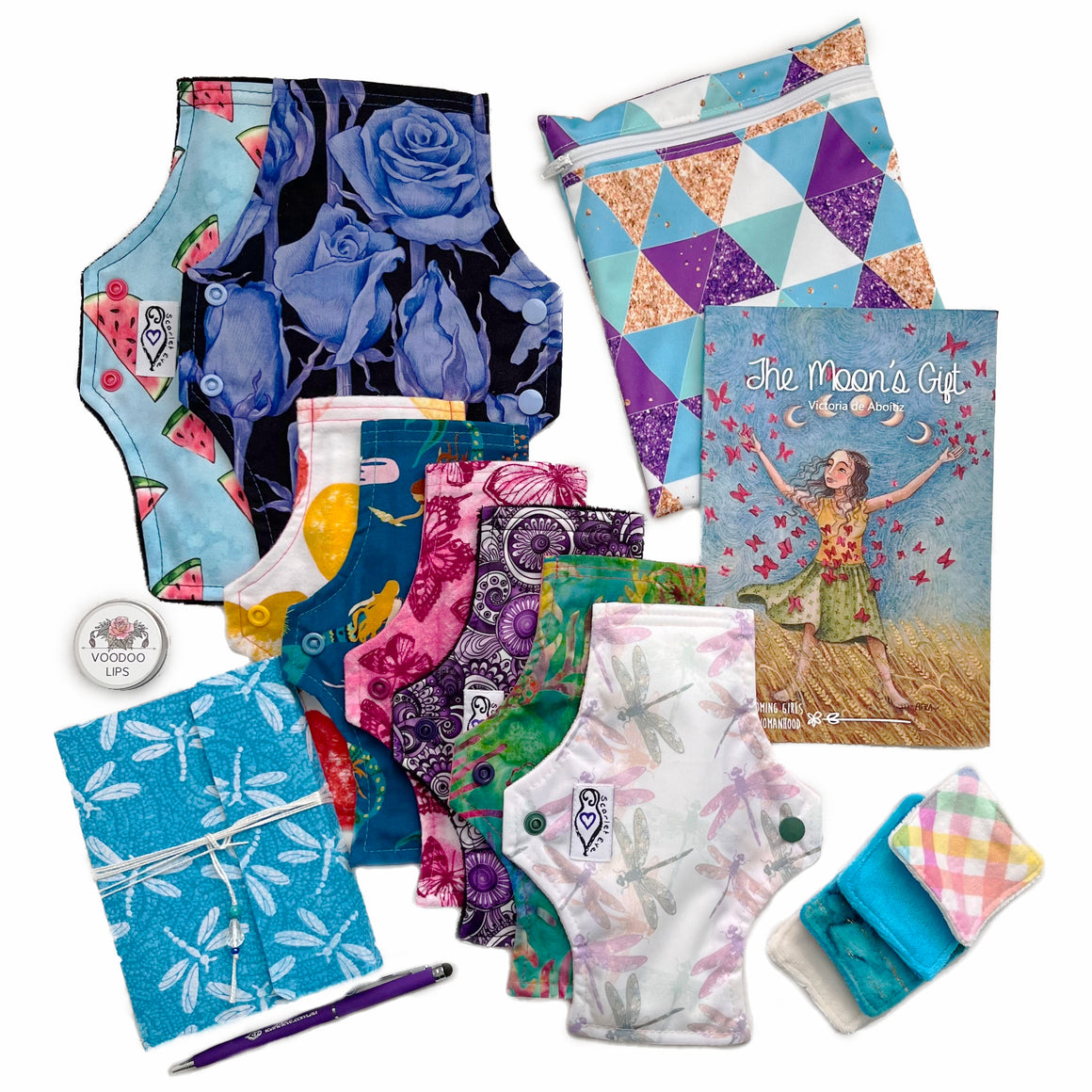 A collection of menstrual products and accessories including pads, a wetbag, journal, pen, lip balm, and face wipes, displayed on a white background.
