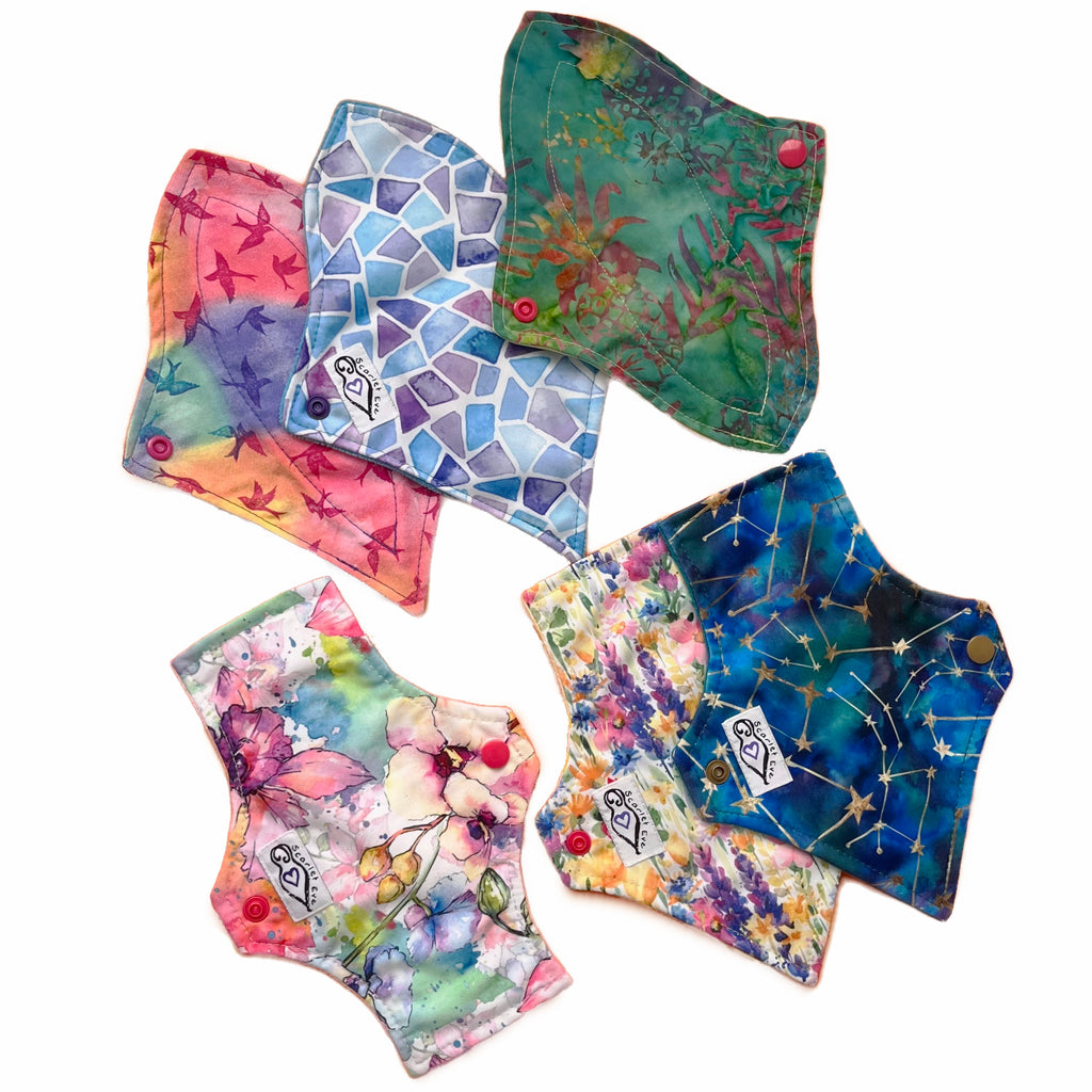 A set of six assorted reusable Mini Pads with various colorful patterns, against a white background. Some Pads are squared at the top and some are curved. Some Pads are tapered at the bottom, one is curved and one is flared.
