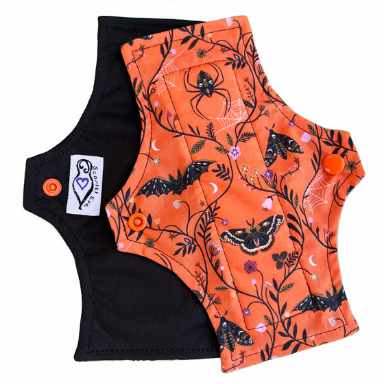 Two reusable mesntrual pads with squared top and flared bottom shape against a white background. The pad on top is made from orange woven cotton with black moths, bats and leaves. The pad on the bottom is made from black PUL fabric and has a white "Scarlet Eve" label.