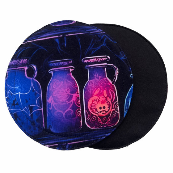 Two reusable breast pads against a white background. The breast pad on top is made from black cotton Lycra with potion bottles in neon colours. The breast pad on the bottom is made from black PUL fabric.