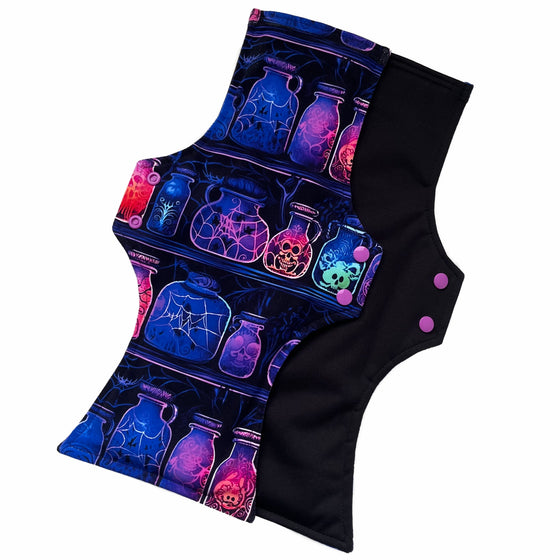 Two reusable menstrual pads with a squared top and flared bottom shape against a white background. The pad on top is made from black cotton Lycra with potion bottles in neon colours. The pad on the bottom is made from black PUL fabric and has a white "Scarlet Eve" label.