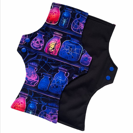Two reusable menstrual pads with a squared top and flared bottom shape against a white background. The pad on top is made from black cotton Lycra with potion bottles in neon colours. The pad on the bottom is made from black PUL fabric and has a white "Scarlet Eve" label.