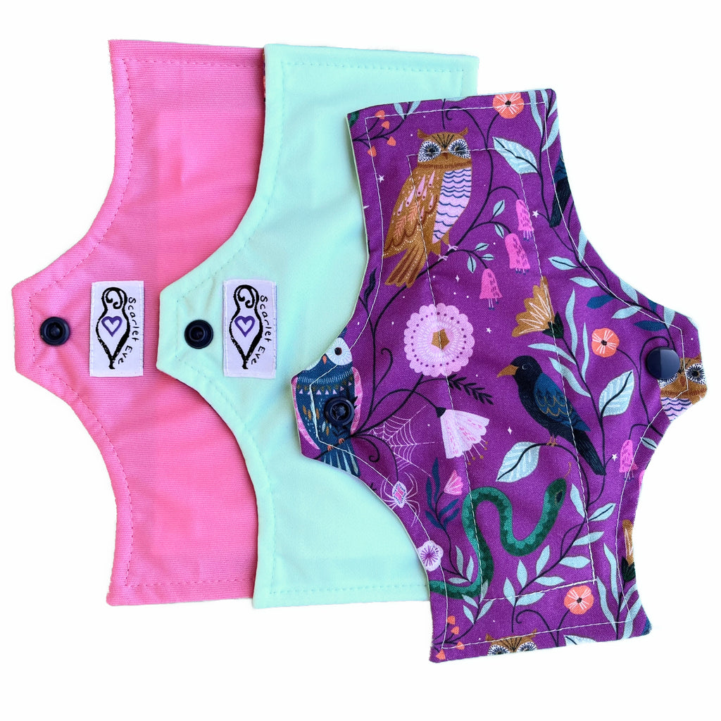 Three reusable menstrual pads with squared top and flared bottom shape, against a white background. The pad on top is made from purple woven cotton with owls, a crow, a snake and leaves. The two pads underneath are made from turquoise PUL fabric with a white "Scarlet Eve" label, and pink PUL fabric with a white "Scarlet Eve" label.