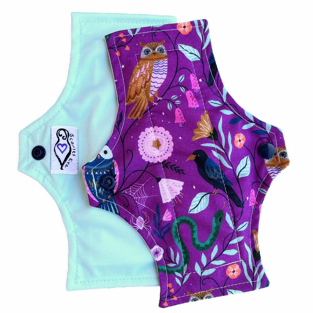 Two reusable menstrual pads with squared top and flared bottom shape, against a white background. The pad on top is made from purple woven cotton with owls, a crow, a snake and leaves. The pad on the bottom is made from turquoise PUL fabric with a white "Scarlet Eve" label.
