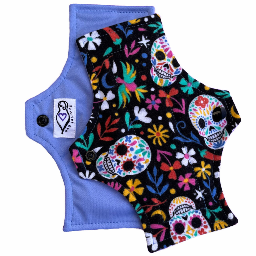 Two reusable menstrual pads with squared top and flared bottom shape, against a white background. The pad on top is made from black flannel fabric with multicoloured sugar skulls and flowers. The pad on the bottom is made from light blue PUL fabric and has a white "Scarlet Eve" label.