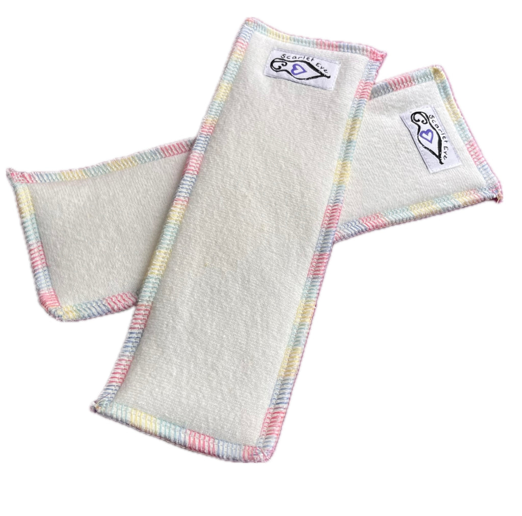 Two rectangular fleece boosters with white "Scarlet Eve" labels, against a white background. The boosters are made from natural fleece with variegated rainbow overlocking.