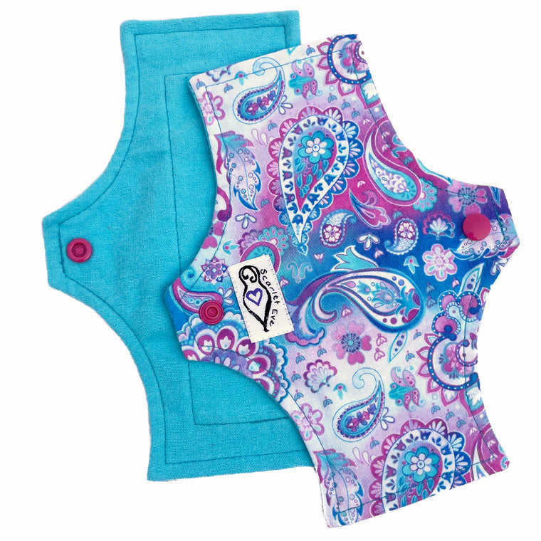 Two reusable panty liners with a squared top and flared bottom shape, against a white background. The pad on the top is made from woven cotton with an aqua blue, purple and white paisley print, and has a white "Scarlet Eve" label. The pad on the bottom is made from aqua blue flannel fabric.