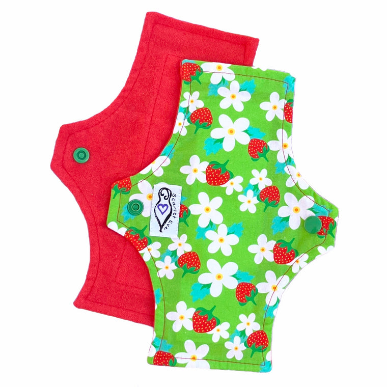 Two reusable panty liners with squared top and flared bottom shape, against a white background. The pad on top is made from bright lime green woven cotton fabric with red strawberries and white flowers, and has a white "Scarlet Eve" label. The pad on the bottom is made from red flannel fabric.