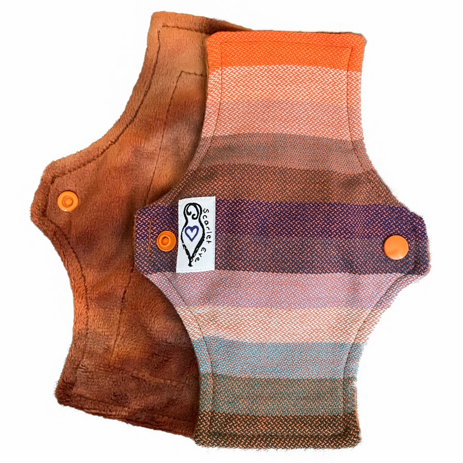 Two reusable panty liners with squared top and flared bottom shape against a white background. The pad on top is made from a babywearing wrap with thick orange, blue and green stripes, and has a white "Scarlet Eve" label. The pad on the bottom is made from caramel brown minky fabric.