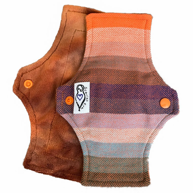 Two reusable panty liners with squared top and flared bottom shape against a white background. The pad on top is made from a babywearing wrap with thick orange, blue and green stripes, and has a white "Scarlet Eve" label. The pad on the bottom is made from caramel brown minky fabric.