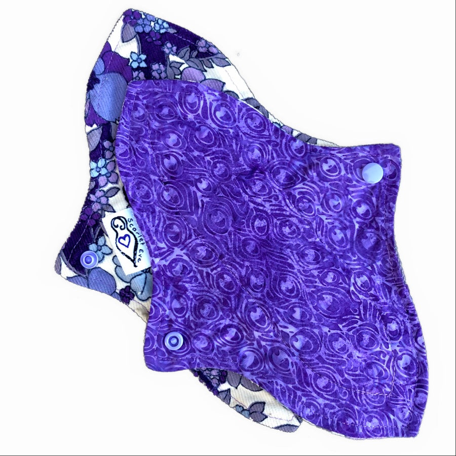 Two reusable menstrual pads with a contoured shape, against a white background. The pad on top is made from purple cotton fabric with batik feathers print. The pad on the bottom is made from purple and white floral corduroy fabric, and has a white "Scarlet Eve" label.