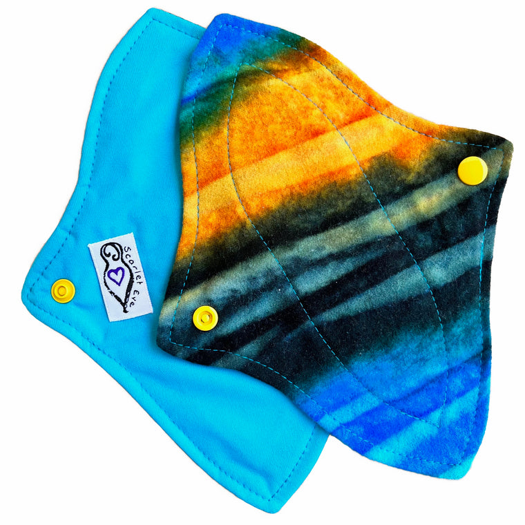 Two reusable menstrual pads with a contoured shape against a white background. The pad on top is made from flannel fabric with batik striations of yellow, aqua and indigo. The pad on the bottom is made from aqua blue PUL fabric.