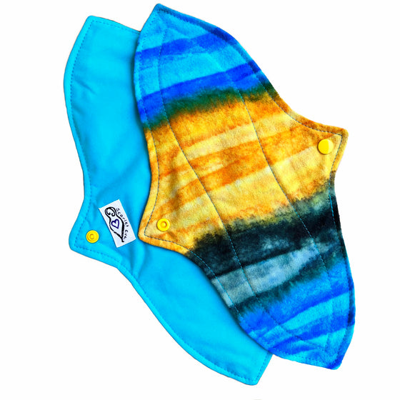 Two reusable menstrual pads with a contoured shape against a white background. The pad on top is made from flannel fabric with batik striations of yellow, aqua and indigo. The pad on the bottom is made from aqua blue PUL fabric.