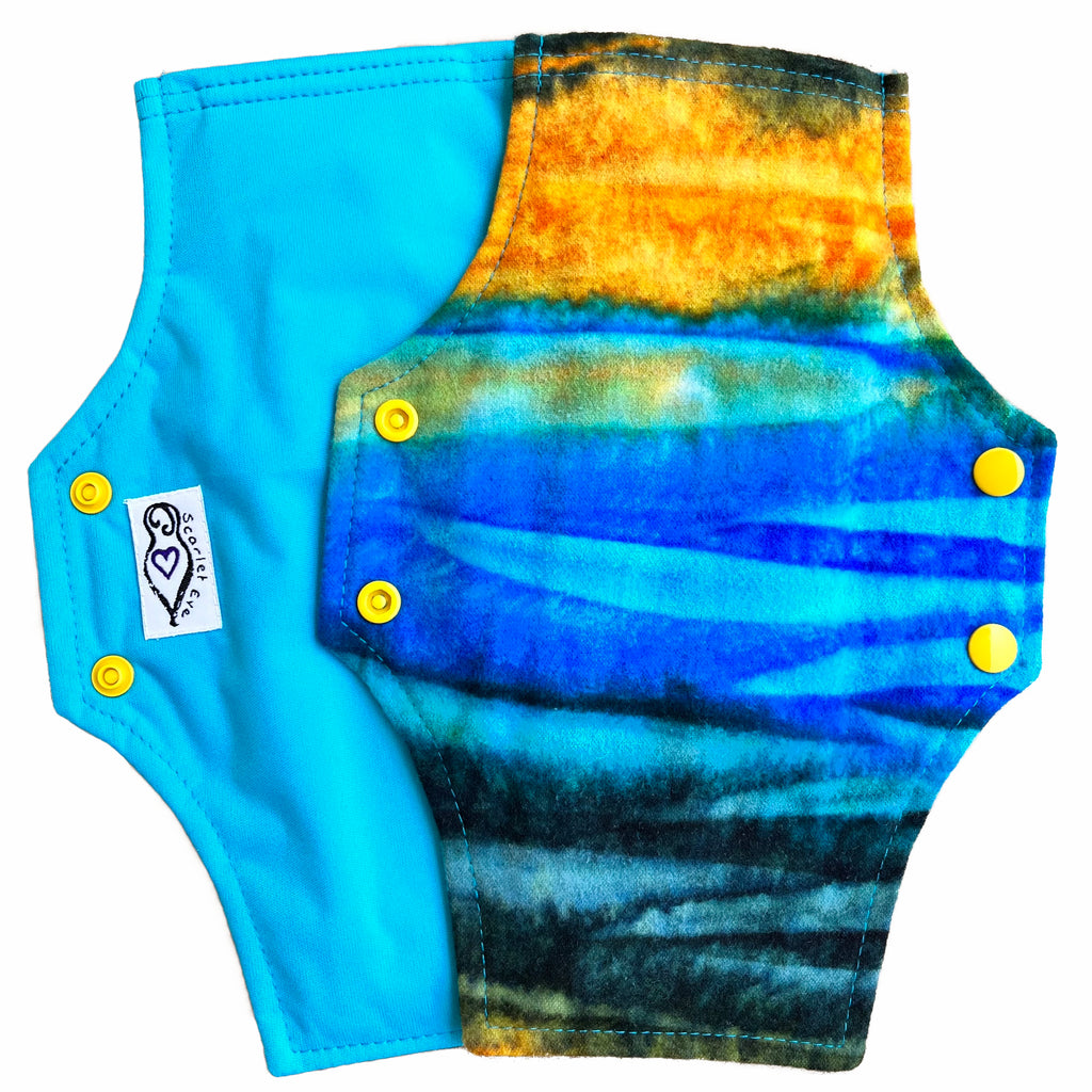 Two reusable menstrual pads with squared top and flared bottom shape against a white background. The pad on top is made from flannel fabric with batik striations of yellow, aqua and indigo. The pad on the bottom is made from aqua blue PUL fabric.